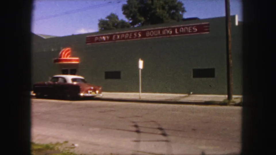 Denver 1958 frame of the bowling alley facade and red signage, with a parked 1950s car along the street in front.