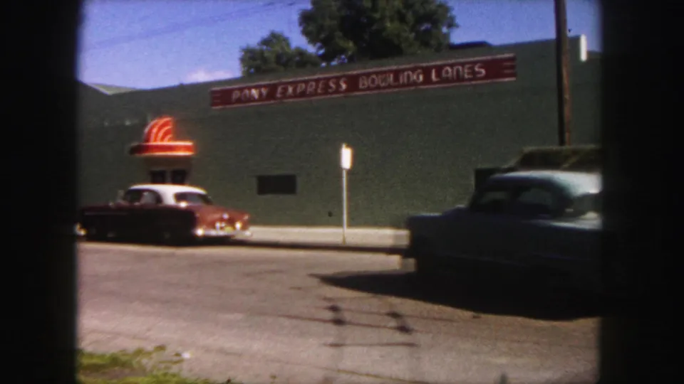 Denver 1958 street frame showing a car passing in the foreground while Pony Express Bowling Lanes and a parked 1950s car remain behind it.