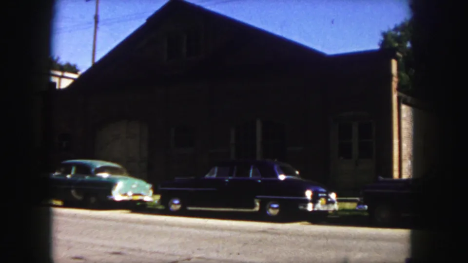Vintage cars are parked along a Denver street in front of a shaded brick building in 1958 home-movie footage.
