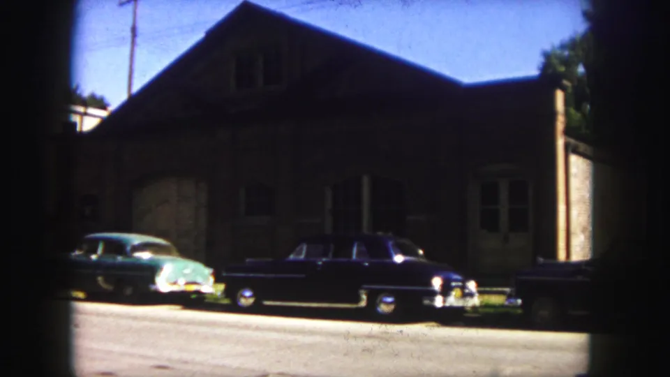 A green car and black sedan are parked near a brick building on a sunlit Denver street in 1958.