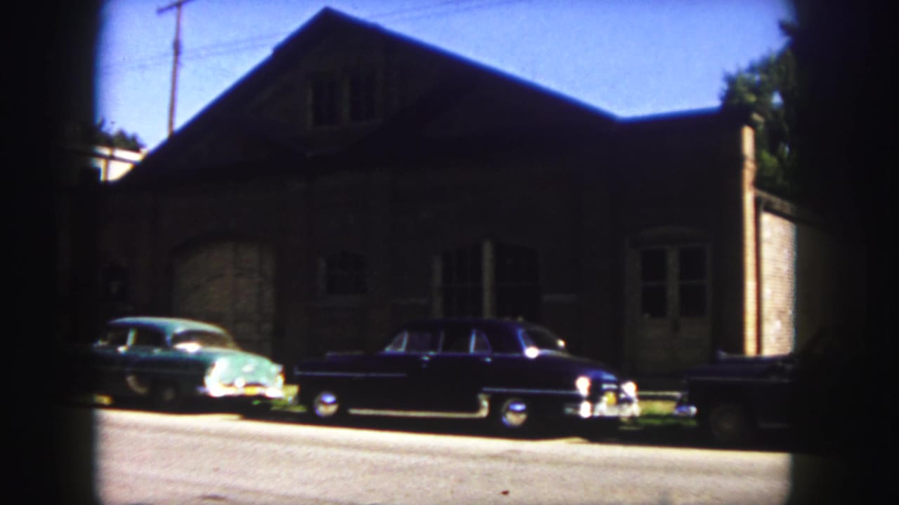 1958 Denver 8mm frame showing a teal car and dark sedan parked in front of a brick building under a blue sky.