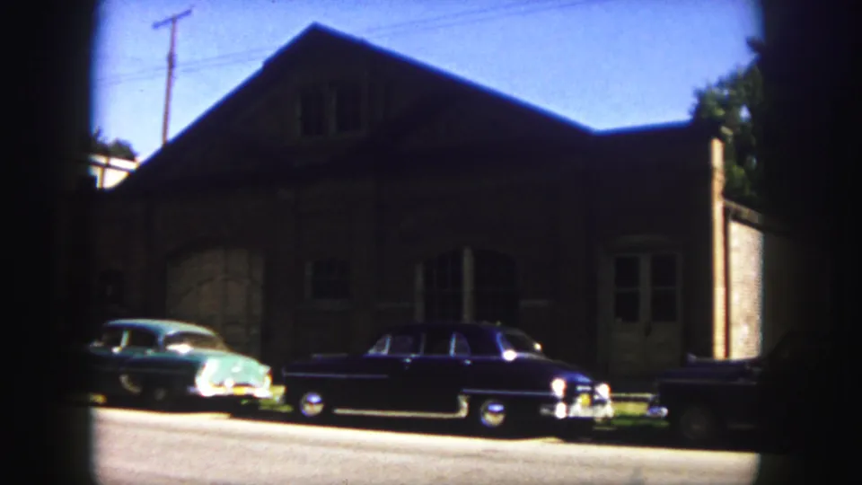 1958 Denver street view with a teal car, dark sedan, brick building, and bright pavement in 8mm color.