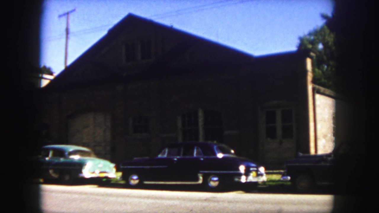 1958 Denver home-movie frame showing two parked classic cars along a street before a large masonry building.
