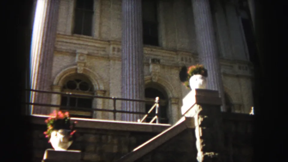 1958 Denver frame showing a closer view of a classical-style building entrance with columns, stone steps, and potted flowers.