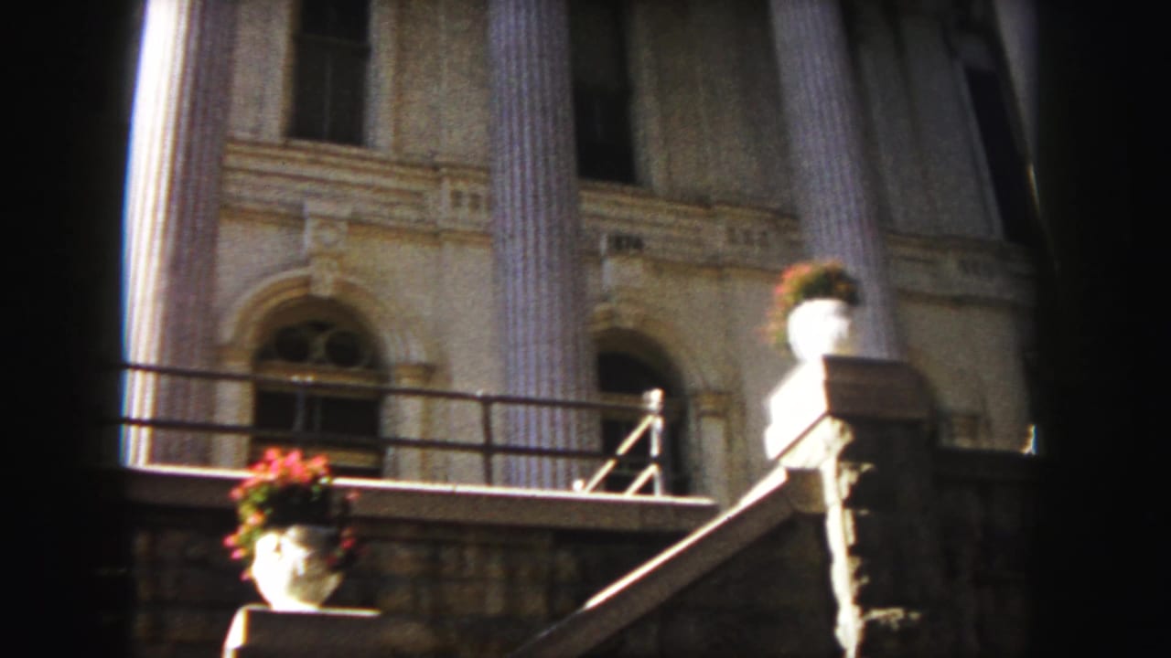 1958 Denver home-movie frame showing a sunlit classical building facade with tall columns, arched windows, stone steps, railings, and flower urns.