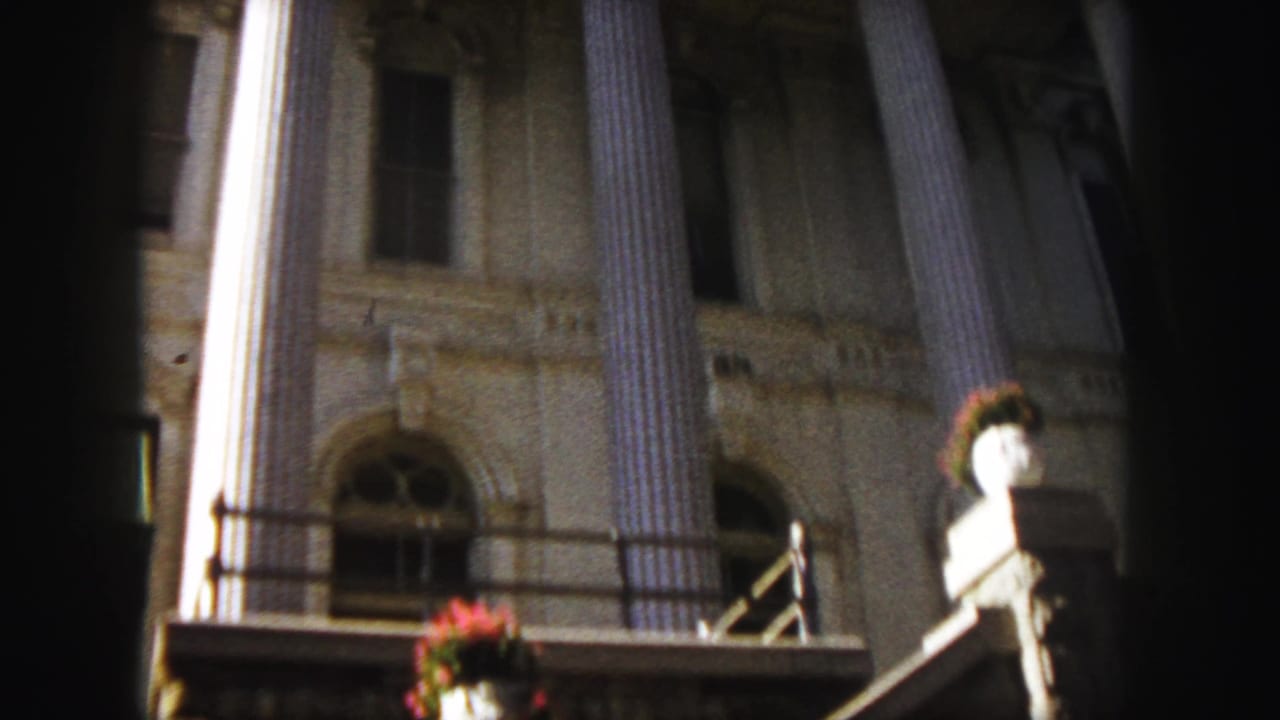 1958 Denver frame from a moving viewpoint showing the classical building's columns, arched window, balcony railing, stone details, and red flowers in a white urn.
