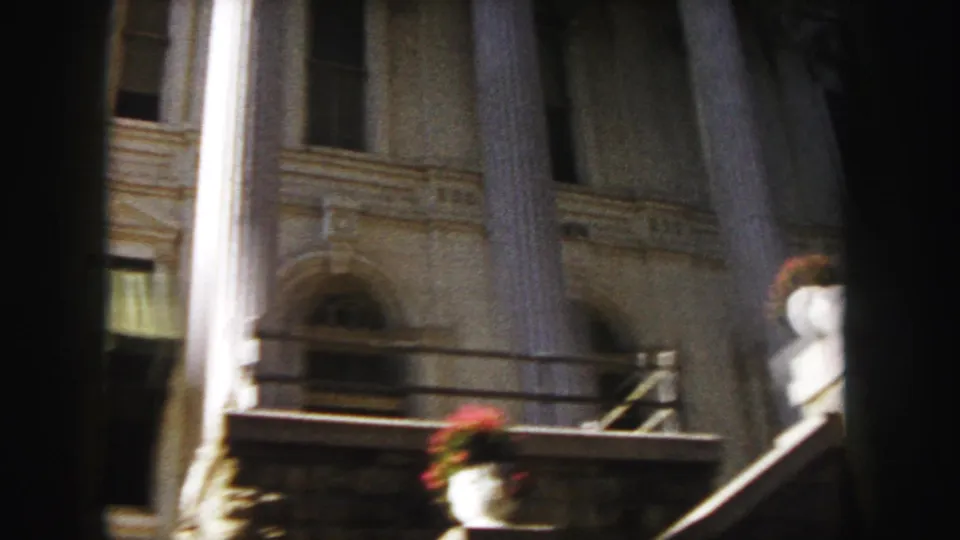 1958 Denver home-movie frame showing an angled, partly shadowed view of a classical stone building with columns, arched windows, steps, railing, and red flower urns.