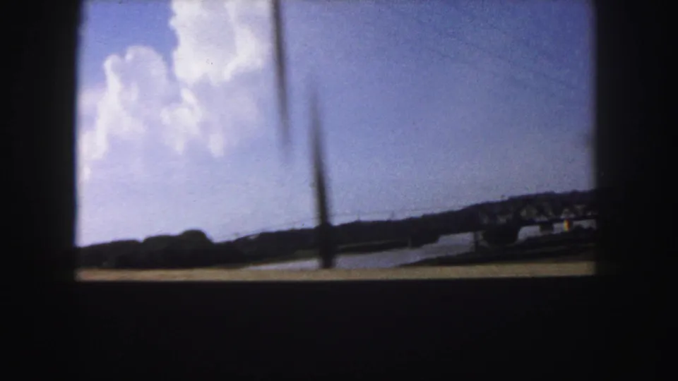 1958 Denver-area travel frame showing a lake or reservoir, distant shoreline, blue sky, and white clouds viewed through a dark vehicle or window frame.