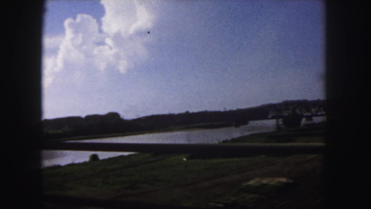 1958 Denver 8mm frame showing a wide river, grassy banks, distant steel truss bridge, and dark foreground vehicle framing.