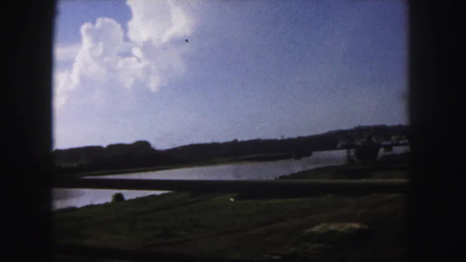 1958 Denver 8mm frame showing a wide river, grassy banks, distant steel truss bridge, and dark foreground vehicle framing.