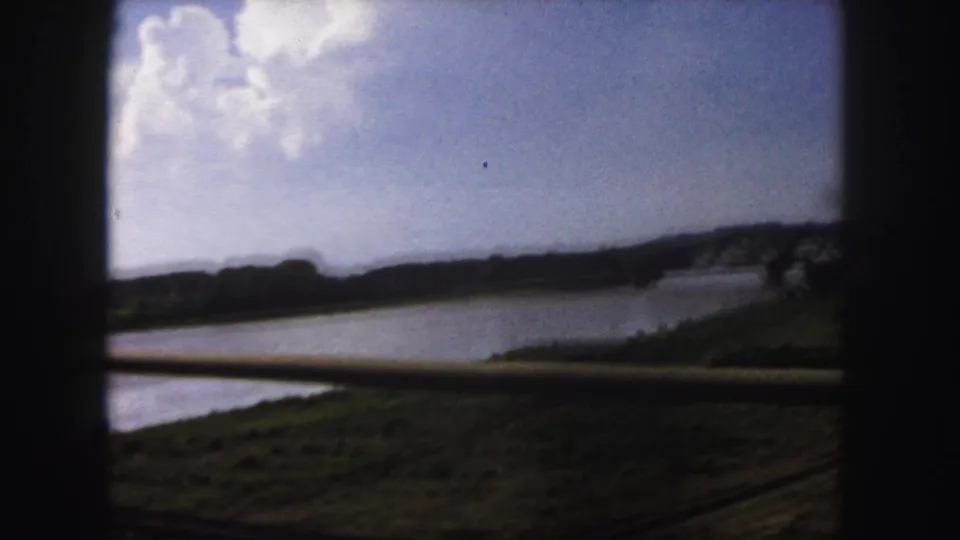 1958 Denver color 8mm frame with the river, green banks, distant bridge, and dark window or railing shapes from a moving vehicle.