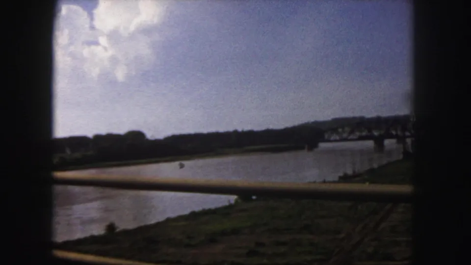 1958 Denver 8mm frame showing the river crossing from a moving vehicle, with a bridge ahead and open banks below the sky.