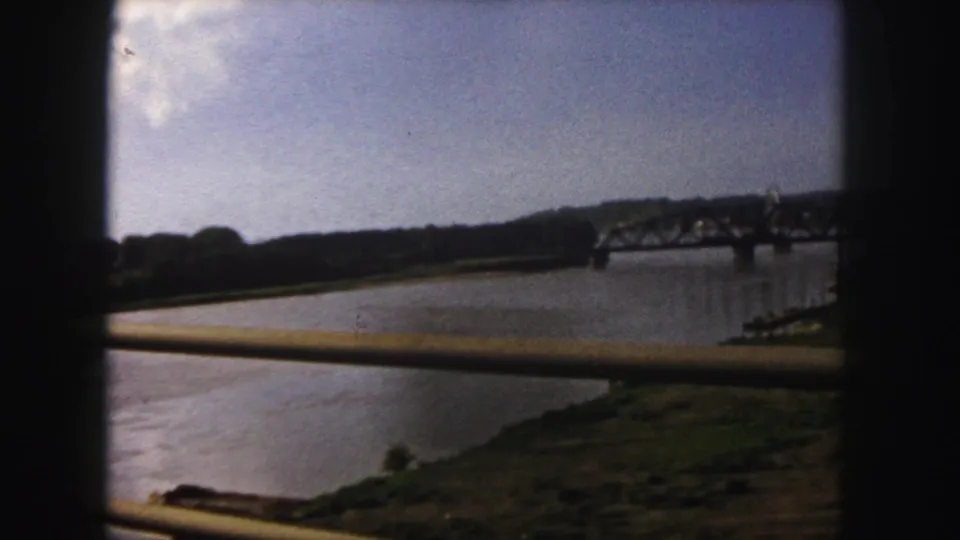 1958 Denver home-movie frame with a riverbank, distant steel truss bridge, and dark foreground framing from a moving crossing.