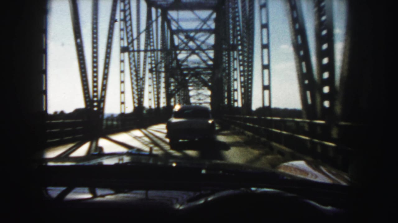 1958 Denver home-movie view through a windshield as a car follows another vehicle across a steel truss bridge.