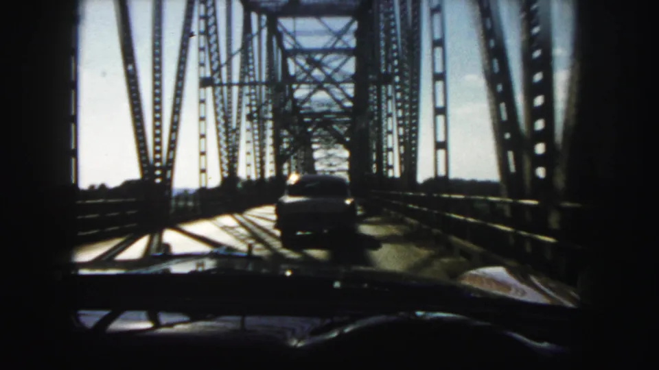 1958 Denver home-movie view through a windshield as a car follows another vehicle across a steel truss bridge.