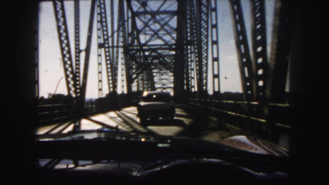 Windshield perspective of 1958 Denver bridge travel, with a car ahead and steel lattice shadows crossing the road.