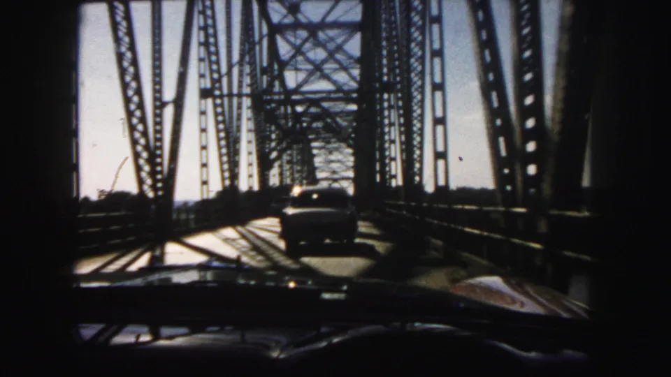 Windshield perspective of 1958 Denver bridge travel, with a car ahead and steel lattice shadows crossing the road.