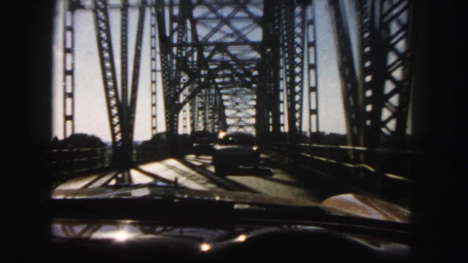 Interior car viewpoint on a Denver steel truss bridge, showing the vehicle ahead framed by dark bridge members and sun glare.