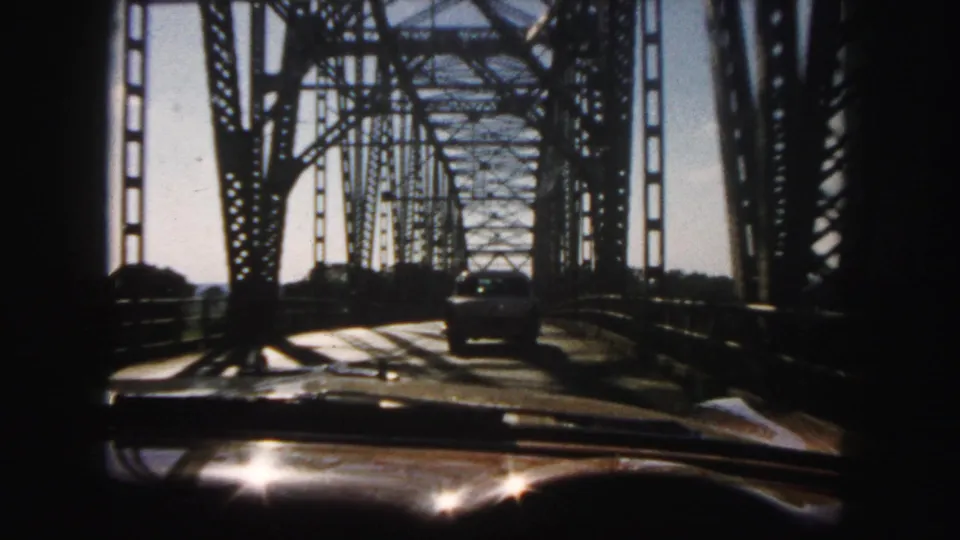 Late-1950s windshield view on a Denver truss bridge, with sunlight flashing on the hood and a car ahead in the lane.