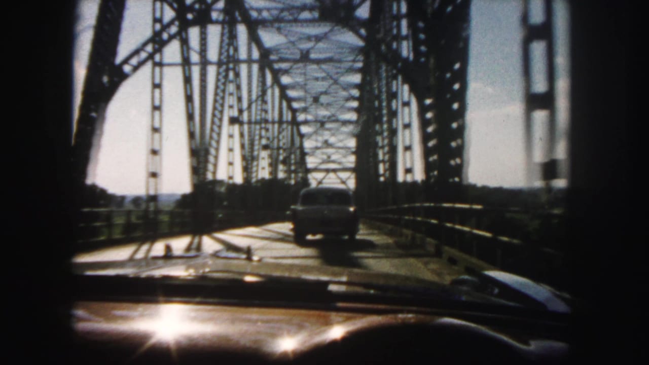 1958 Denver-area home-movie frame showing a car moving across a steel truss bridge, filmed through the windshield from inside another vehicle.