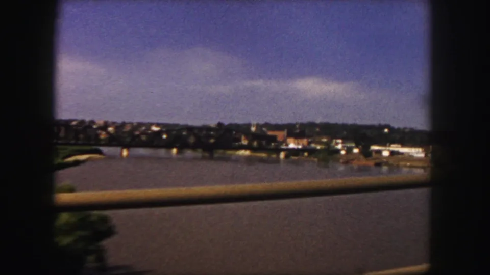 1958 Denver-area travel footage shifts from the bridge to a view across water toward a distant shoreline, with a foreground railing from the moving car.
