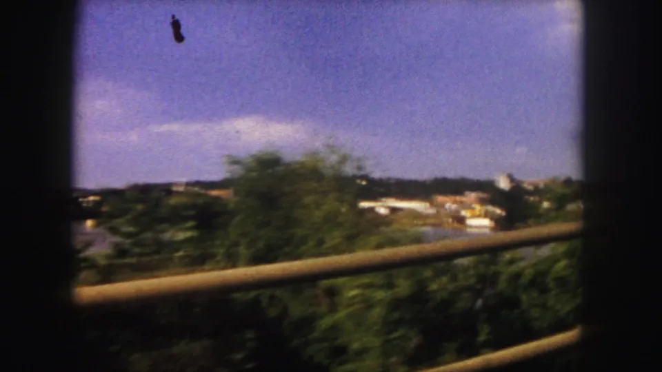 The 1958 home-movie view passes trees and a rail beside water, with distant buildings visible across the shoreline.