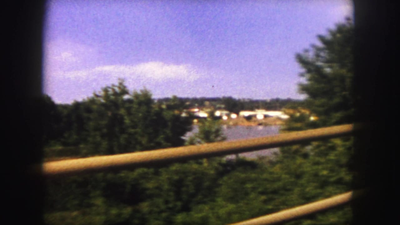 1958 8mm Denver-area travel view with green trees, a wooden railing, distant low buildings, and blue sky.