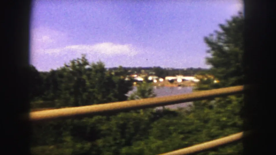 1958 8mm Denver-area travel view with green trees, a wooden railing, distant low buildings, and blue sky.