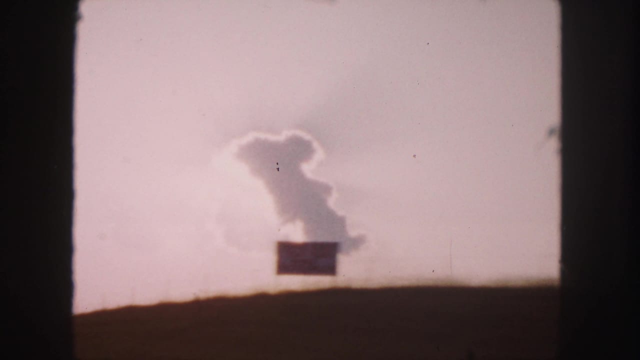 1958 8mm road-trip frame showing a pale sky, distinctive cloud shape, dark horizon, and small distant roadside sign.
