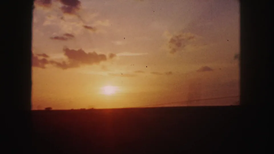 1958 8mm road-trip frame showing a warm sunset, scattered clouds, and a dark silhouetted horizon.