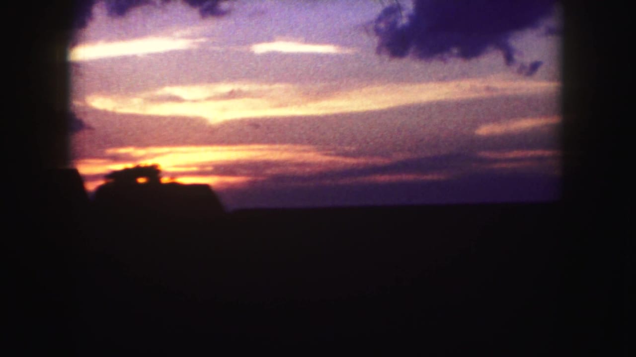 1958 8mm Denver metadata frame showing a purple-orange sunset beyond dark silhouetted land, viewed from inside a moving car.