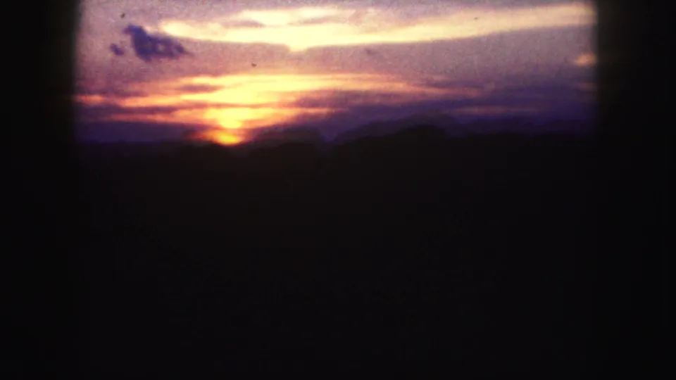 A low sun glows through cloud bands over a dark silhouetted horizon in this 1958 Denver 8mm home-movie frame.
