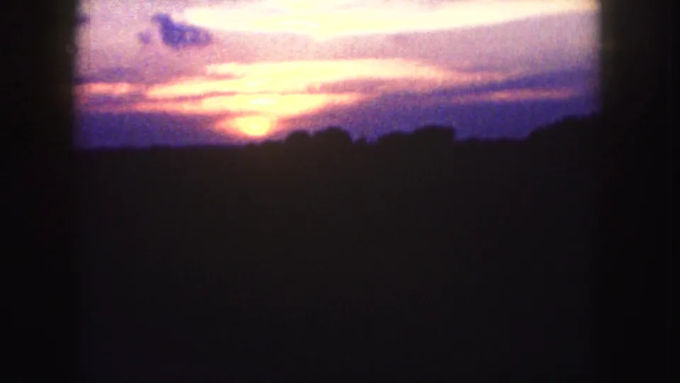 Soft 8mm frame of an orange sunset fading behind a dark tree or hill silhouette, with magenta and purple sky tones.