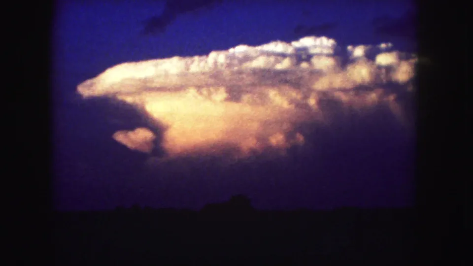 1958 Colorado home-movie frame with a wide illuminated cloud band over a low, dark tree line.