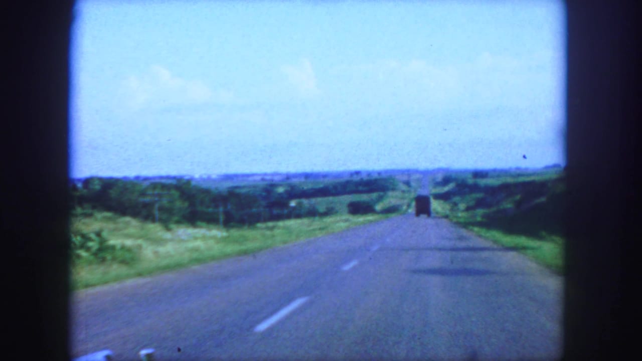 1958 8mm view from a car on a paved road near Denver, with green fields, blue sky, and a distant truck ahead.