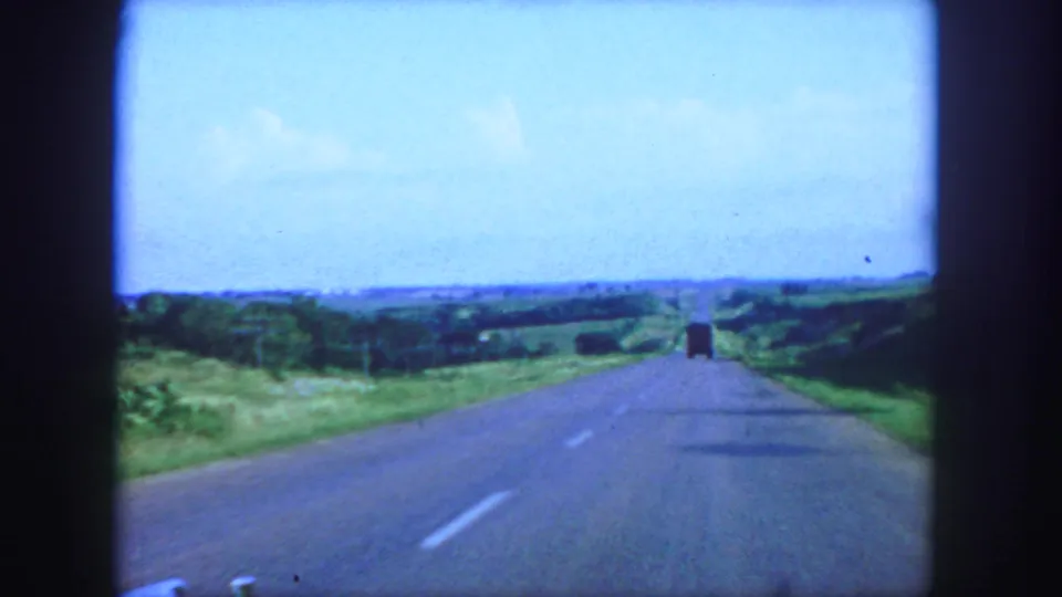 1958 8mm view from a car on a paved road near Denver, with green fields, blue sky, and a distant truck ahead.