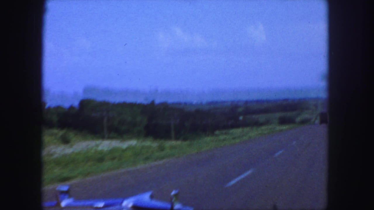 Curving paved road from a car with the hood visible and fields beyond.