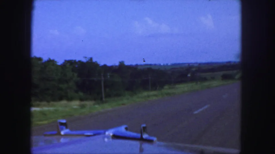 1958 home-movie view from a moving car near Denver, showing the hood, paved road, trees, fields, and a hazy blue sky.