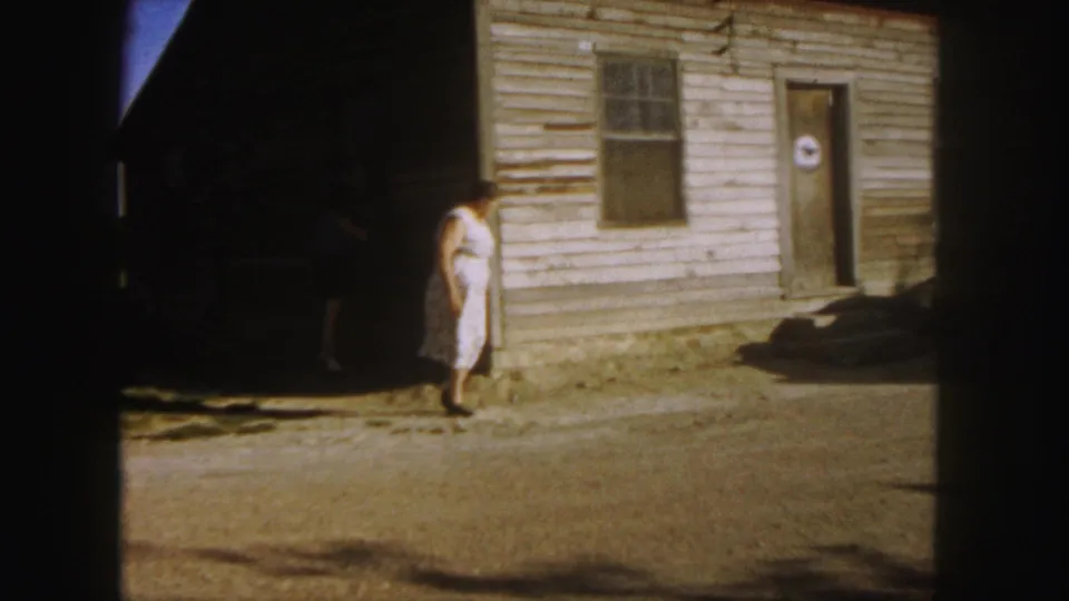 A woman in a white dress stands beside a weathered wooden building in 1958 8mm footage from the Denver metadata set.
