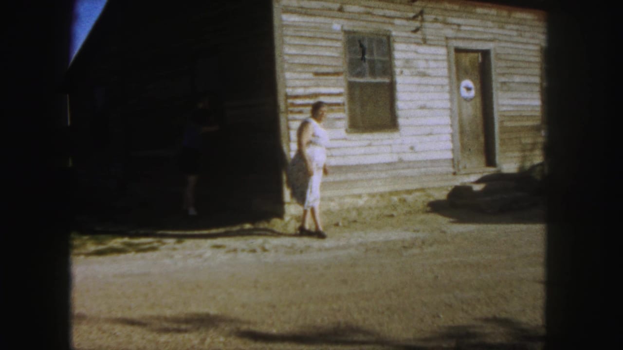 In 1958 Denver home-movie footage, a woman in a white dress stands near a clapboard building while another person is partly visible in shade.