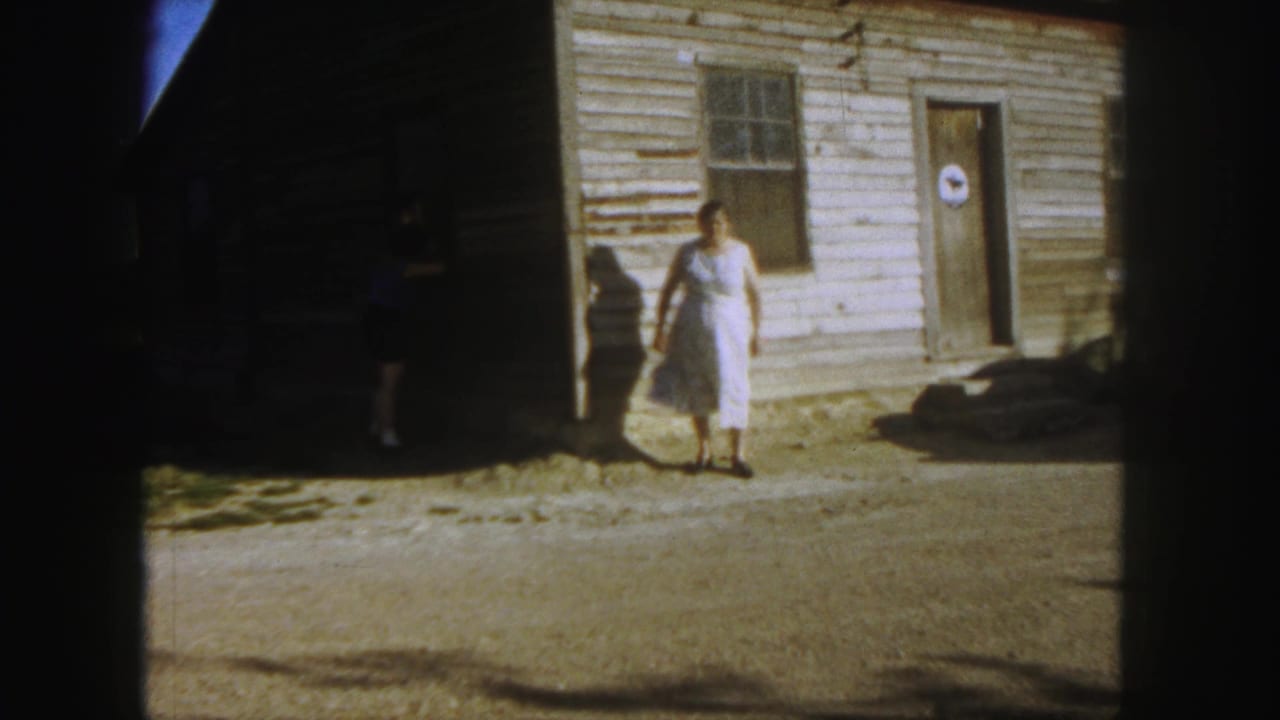 Woman in a white dress walks across a dirt yard in front of a clapboard building.