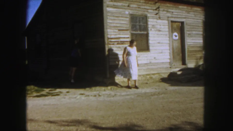 The woman continues across the sunlit yard in 1958 Denver footage, passing the clapboard wall as a second person remains in dark shade.