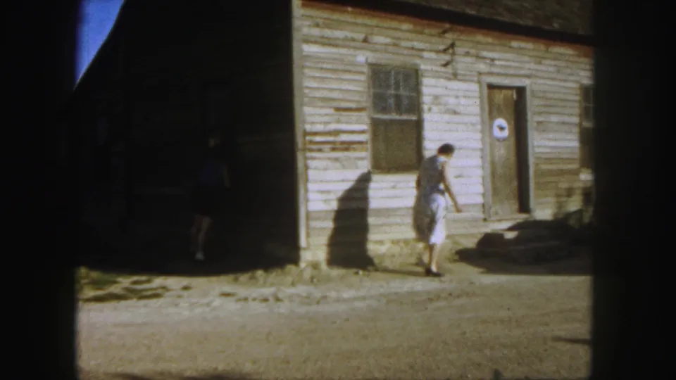At the end of the clip, the woman leans or steps forward near the clapboard building, with the round door sign and heavy shadows still visible.