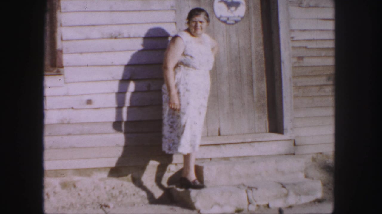 In 1958 Denver home-movie footage, an older woman stands on stone steps by a weathered wooden doorway in bright sun.