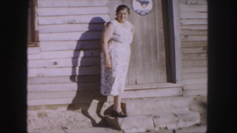 In 1958 Denver home-movie footage, an older woman stands on stone steps by a weathered wooden doorway in bright sun.