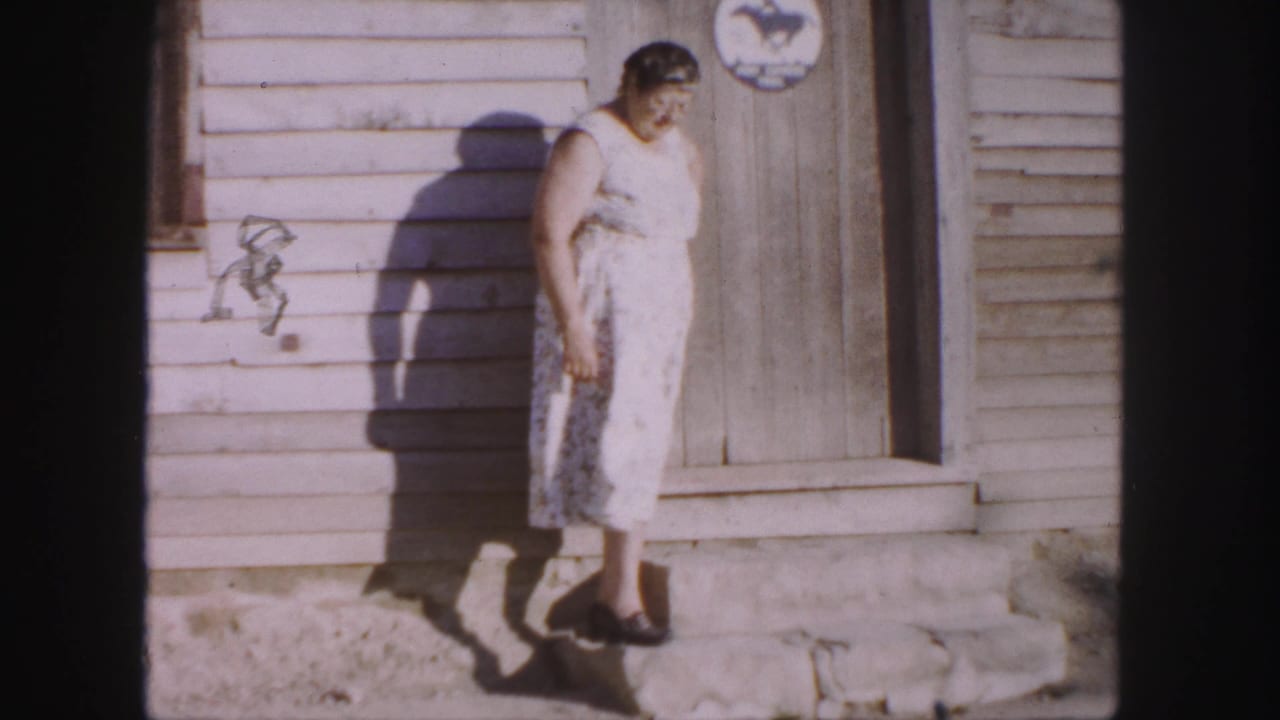 Older woman looks down while standing beside a wooden door and wall decoration.