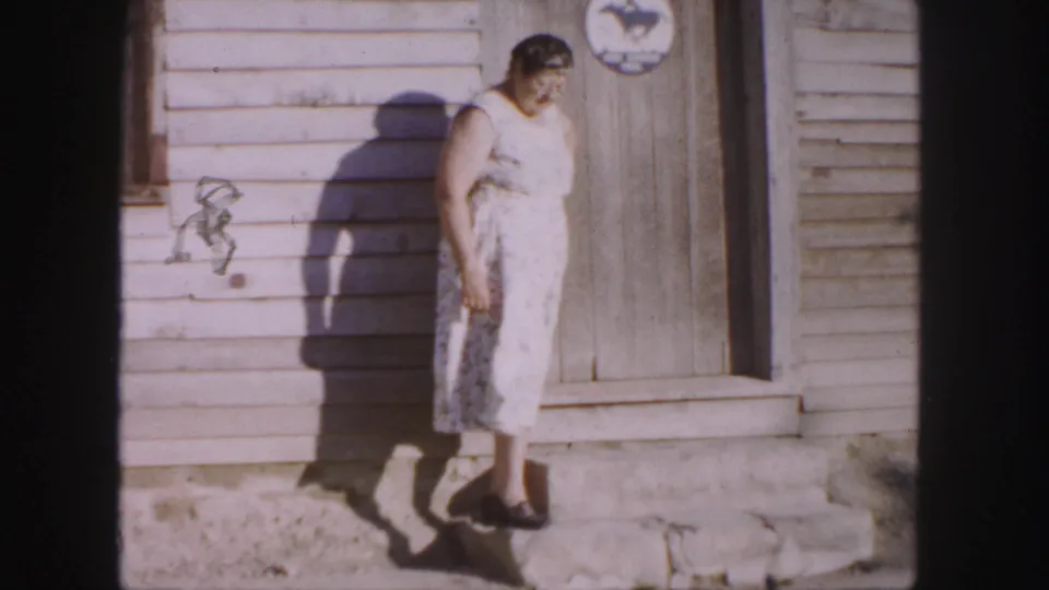 The older woman looks down on the stone step outside a Denver wooden building, with her shadow falling across the siding.