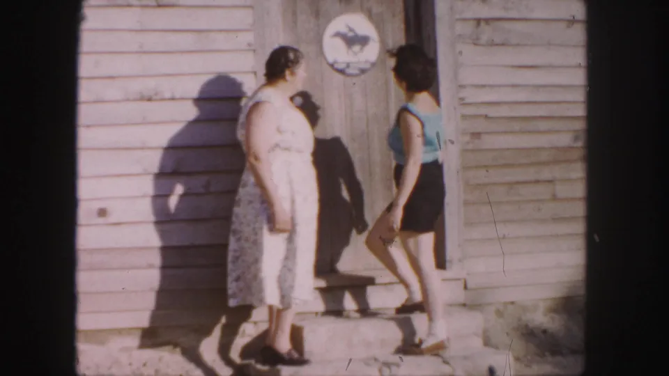 The younger woman in a blue top steps toward the older woman near a wooden doorway in the Denver home-movie scene.