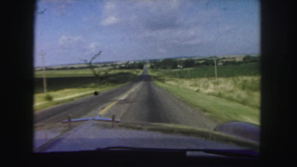 1958 8mm view from inside a car driving along a straight rural road near Denver, with fields and utility poles visible.