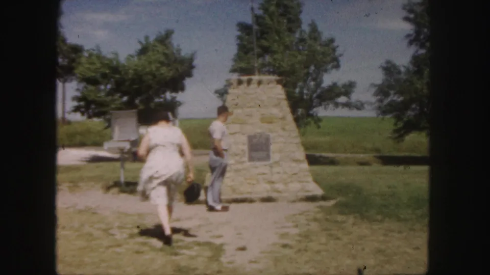 Two adult visitors approach a stone monument with a plaque in a grassy area during the 1958 Denver-area travel footage.
